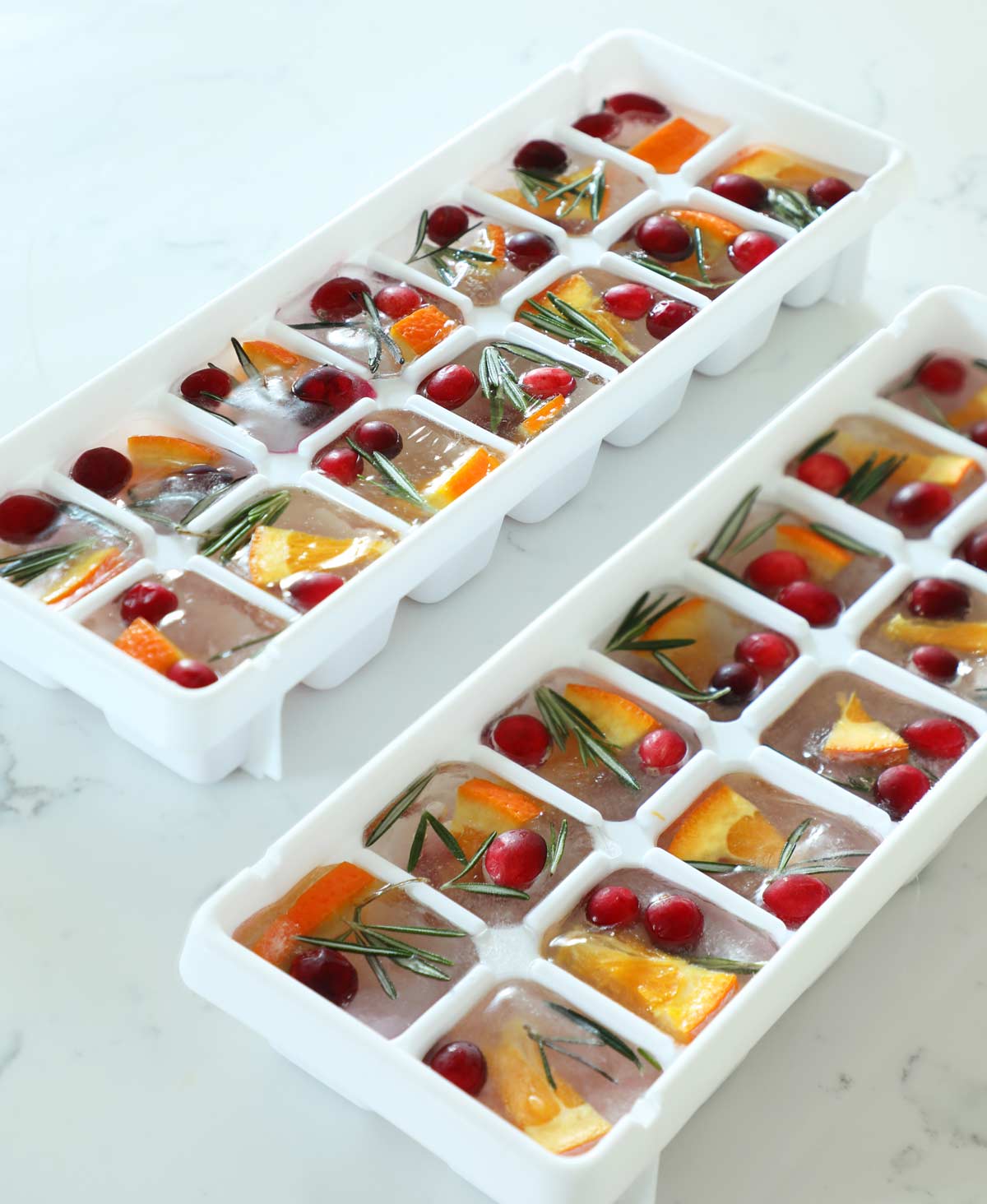 Two ice cube trays filled with infused ice cubes for Christmas on a white countetop.