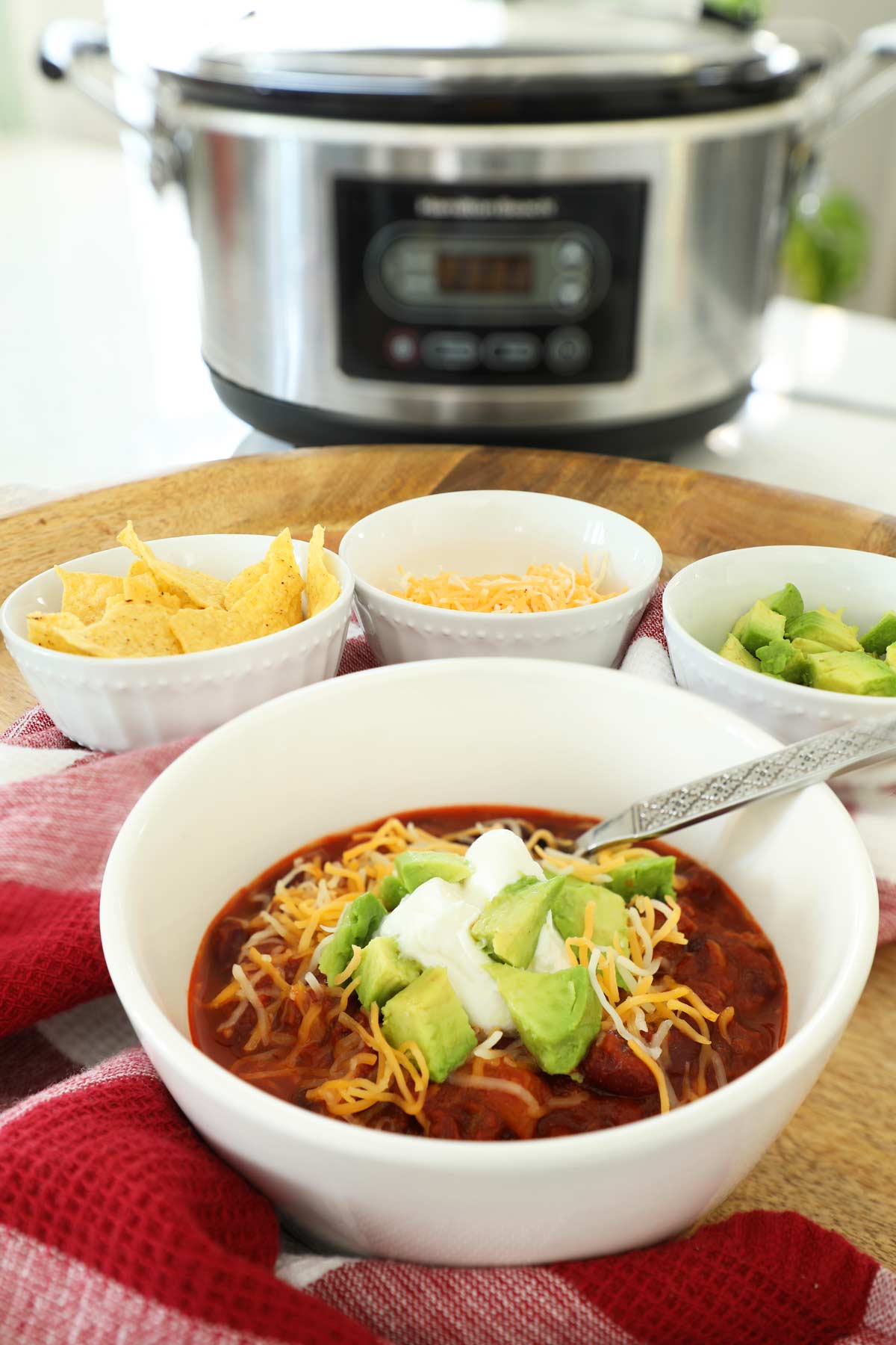 Sausage Chili in a white bowl topped with cheddar sour cream and avocado on a wooden tray with a slow cooker in background.