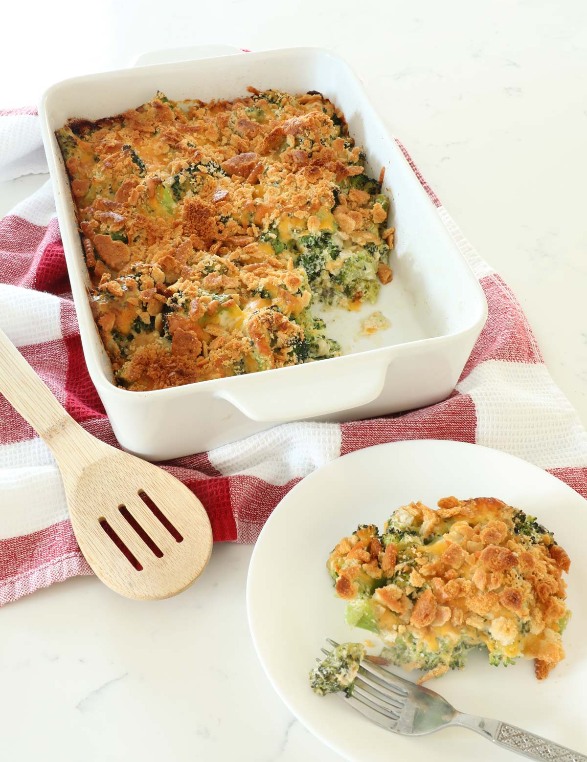 Broccoli Casserole in a white baking dish with a scoop of casserole on a white plate on a red and white kitchen towel. 