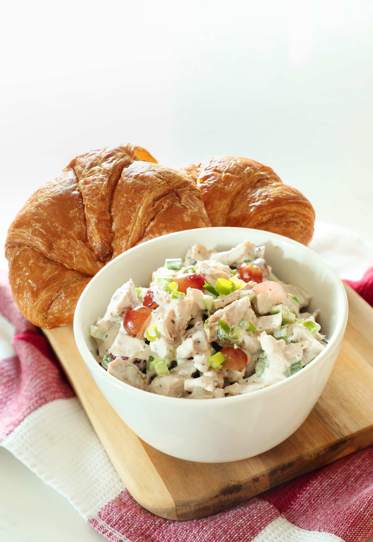 White bowl filled with turkey salad on a wooden cutting board with two croissants on a kitchen counter. 