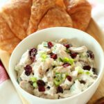 Turkey salad with cranberries in a white bowl next to a couple croissants sit on a wooden board on top of the kitchen counter.