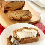 Sliced zucchini bread sits on a wooden board on top of the kitchen counter, one slice with a butter smear sits on a white plate accompanied by a fork.