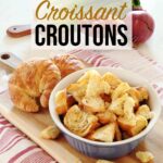 Croissant Croutons gathered in a blue bowl resting on a wooden board accompanied by a couple unsliced croissants, red onion, tomatoes and lettuce in the background.