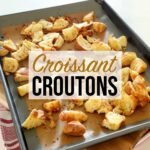 Freshly baked croissant croutons resting on a metal baking sheet on a wooden board on top of the kitchen counter.