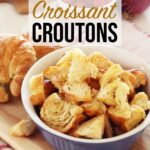 Croissant Croutons gathered in a blue bowl resting on a wooden board accompanied by a couple unsliced croissants, red onion, tomatoes and lettuce in the background.