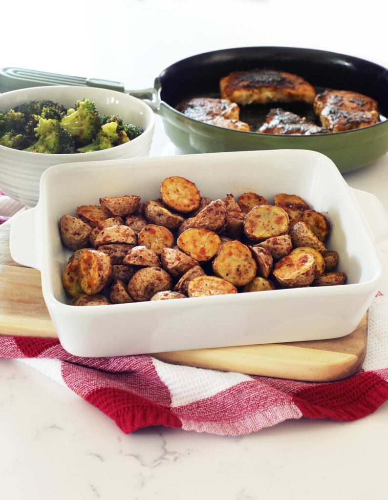 A white baking dish of AirFryer Baby Potatoes on a cutting board with cast iron pork chops and broccoli.