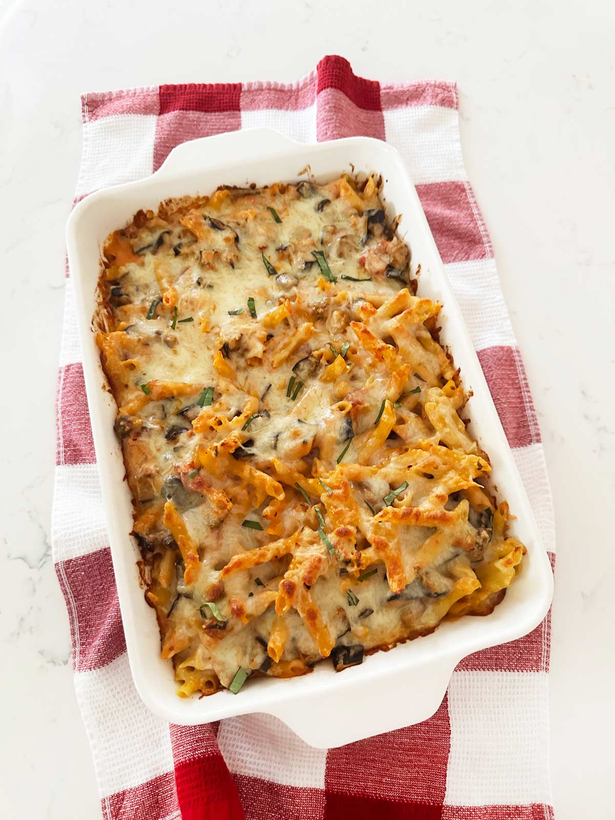 Baked Penne with eggplant in a casserole dish on a red and white kitchen towel on a white kitchen countertop.
