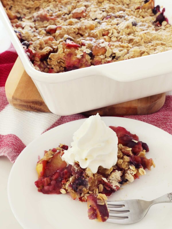 Fresh baked Fruit Crisp with peaches in a white baking dish on top of the kitchen counter with a portion of fruit crisp on a white plate and whipped cream on top.