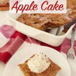 Freshly baked apple cake in a white baking dish atop the kitchen counter, on a red towel with one slice of apple cake on a white plate in the foreground with whipped cream on top.