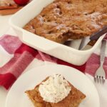 Freshly baked apple cake in a white baking dish atop the kitchen counter, on a red towel with one slice of apple cake on a white plate in the foreground with whipped cream on top.