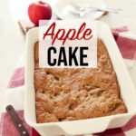 Freshly baked apple cake in a white baking dish atop the kitchen counter, on a red towel with a spatula in the foreground, and an apple, plates and forks In the background.