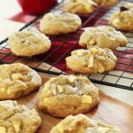 Apple cookies rest on a wooden board and a baking rack all on top of the kitchen counter.