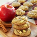 Apple cookies are stacked 3 high on a wooden board with an apple and cinnamon sticks off to the side, while in the background apple cookies cool on a baking rack.