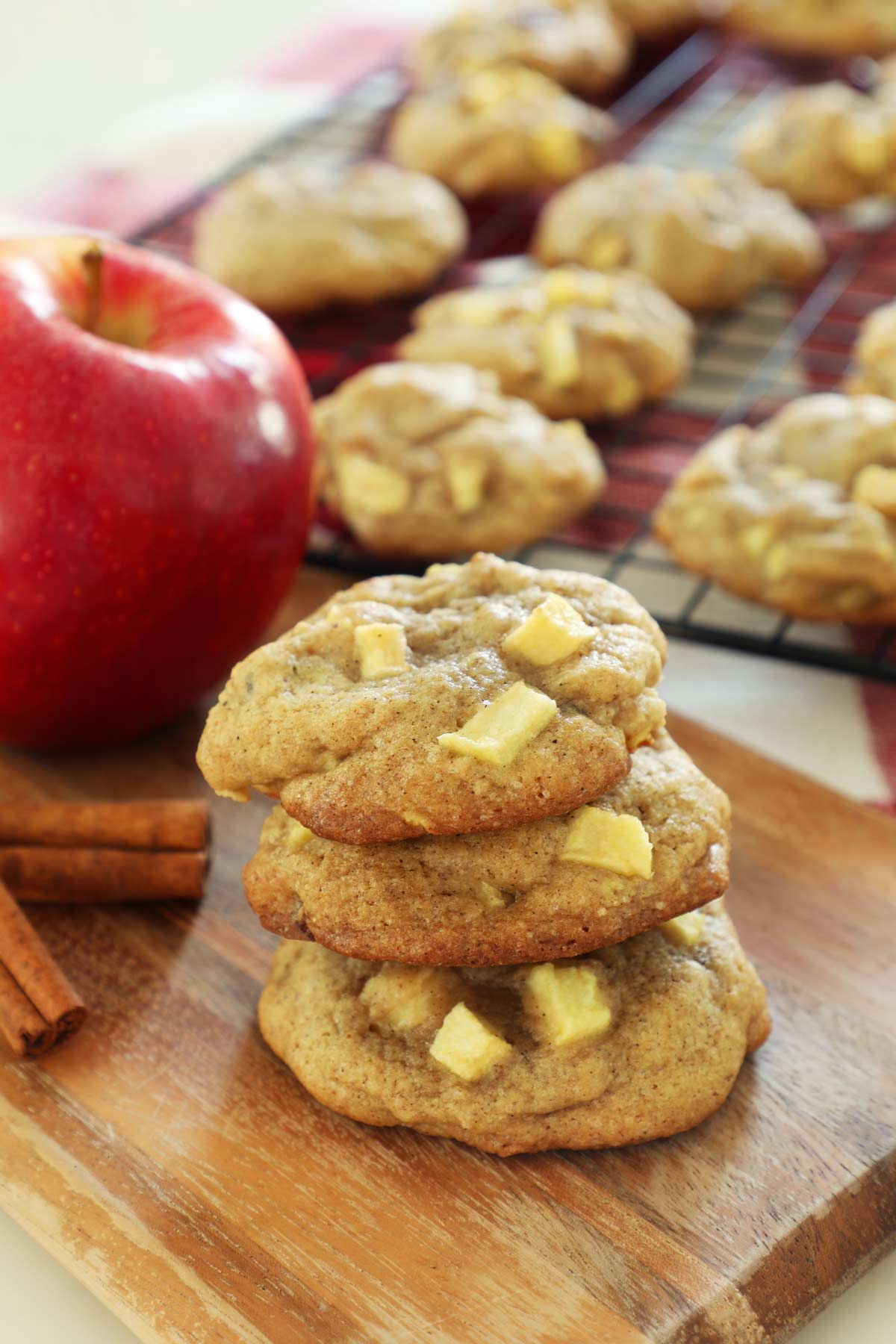 Three apple cookies stacked on a cutting board with a cookie cooling rack in the background with cinnamon sticks and an apple.