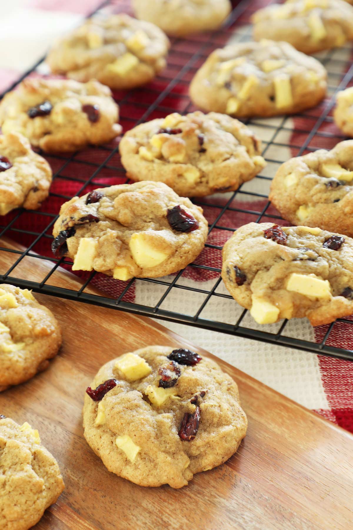 Apple cranberry cookies cooking on a baking rack, one cookie sits on a wooden board.