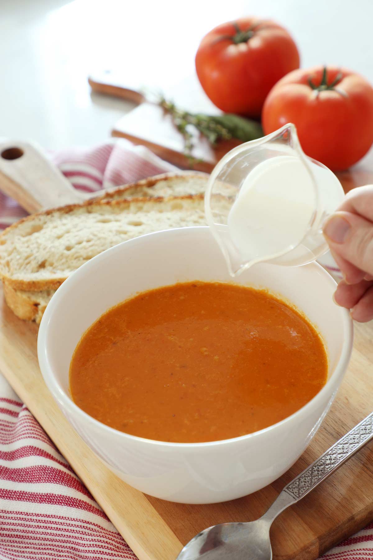 A white bowl filled with tomato and roasted red pepper soup with cream being poured in.