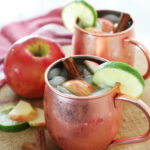 Two apple cider mules on a wooden cutting board with sliced apples and limes.