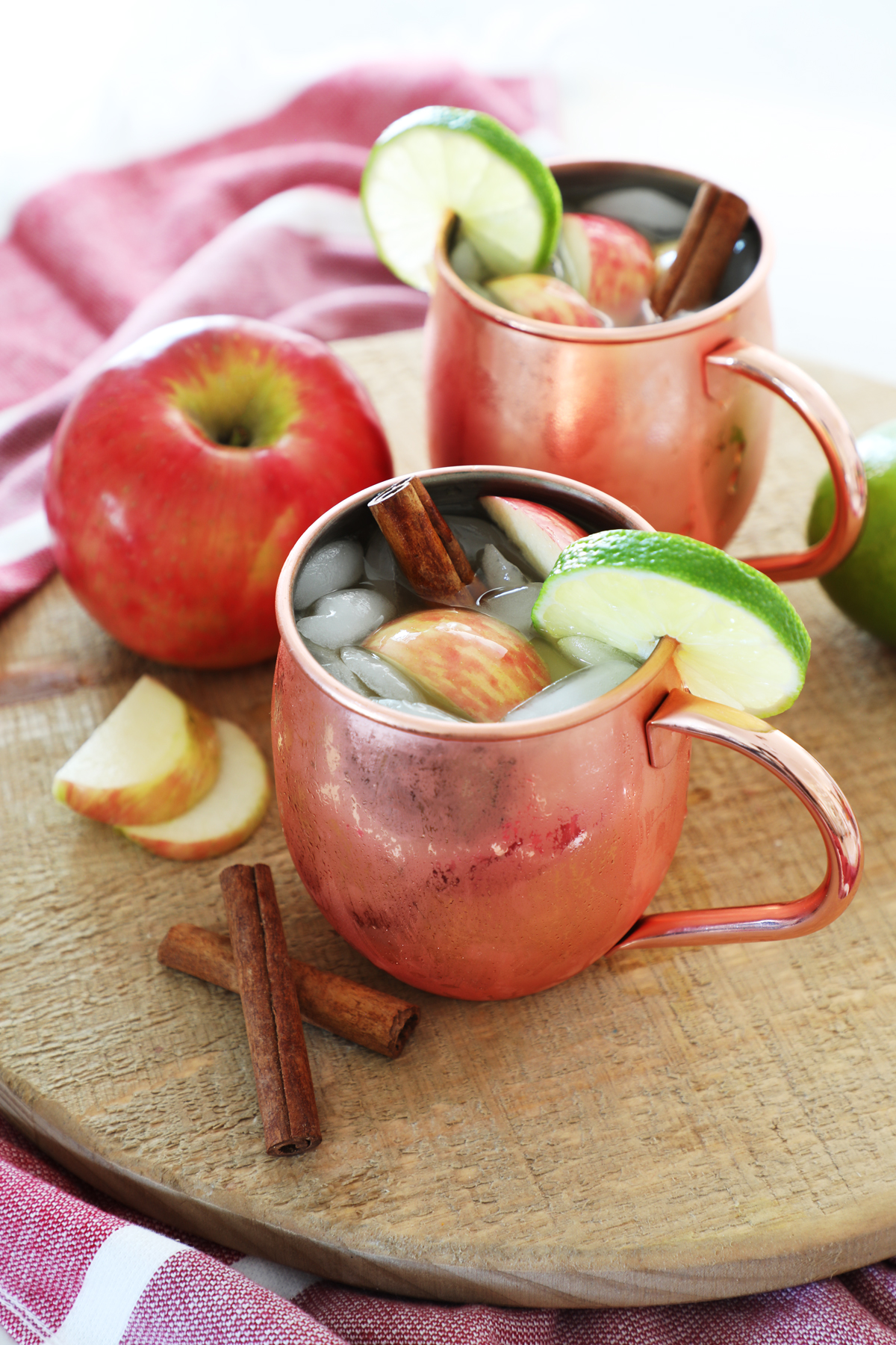 Two copper mugs filled with apple cider mules, ice, cinnamon sticks, apple slices and lime slices on a cutting board.
