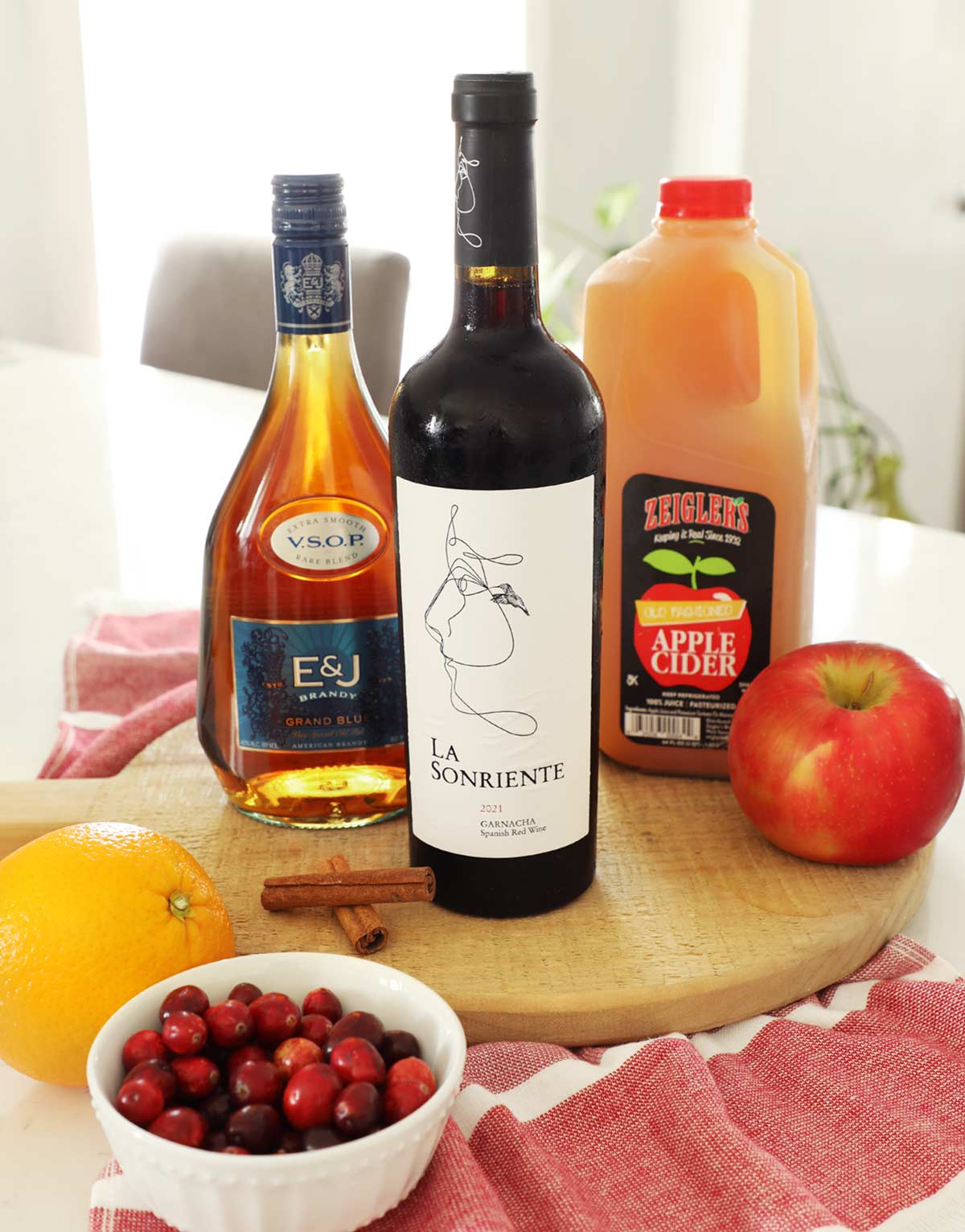 Cutting board on a kitchen counter with fall sangria ingredients brandy, red wine, apple cider, apples, cinnamon sticks, orange and cranberries in a small white bowl.