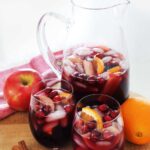 Pitcher of red apple cider sangria on a wooden cutting board with two glasses.