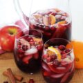 A pitcher of fall sangria with two full wine glasses on a wooden cutting board.