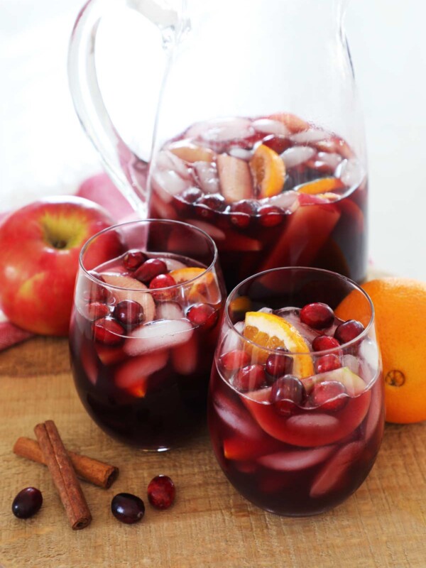 A pitcher of fall sangria with two full wine glasses on a wooden cutting board.