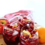 3 wine glasses filled with red apple sangria on a kitchen counter with an apple and orange on the counter.
