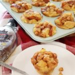 Baked french toast muffins in a light blow muffin tin on a red kitchen towel with a small white plate with a muffin on it.