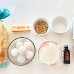 Brioche, eggs, butter, brown sugar, milk, sugar, vanilla extract, cinnamon and salt laid down on a kitchen counter.