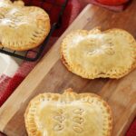 Apple Hand Pies shaped like apples resting on cooling rack, wooden board and red towel on kitchen counter.