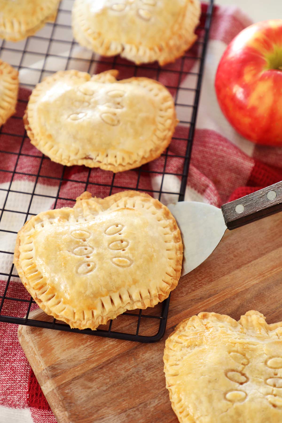 Apple Hand Pies shaped like apples resting on cooling rack, wooden board and red towel on kitchen counter with red apple in background.