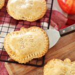 Apple Hand Pies shaped like apples resting on cooling rack, wooden board and red towel on kitchen counter, apple In background.