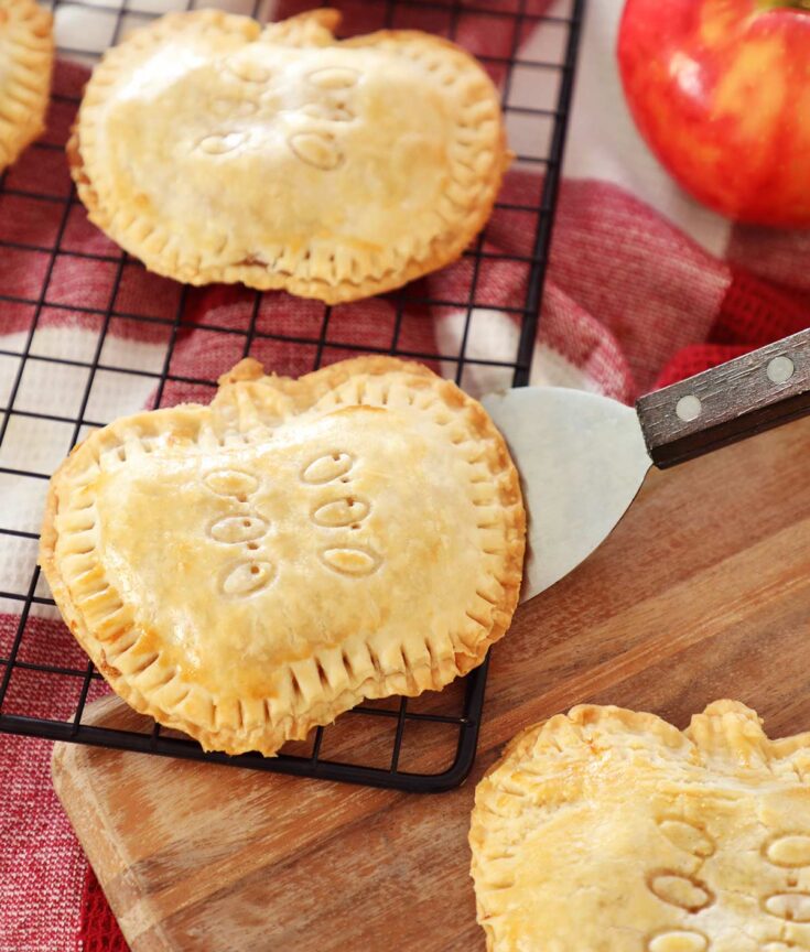 Apple Hand Pies shaped like apples resting on cooling rack, wooden board and red towel on kitchen counter, apple In background.