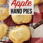 Apple Hand Pies shaped like apples resting on cooling rack, wooden board and red towel on kitchen counter with an apple.