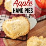Apple Hand Pies shaped like apples resting on cooling rack, wooden board and red towel on kitchen counter.