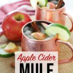 Apple Cider Mule on kitchen counter, sitting atop a red towel and wooden Board with apples, limes and cinnamon stick garnish.