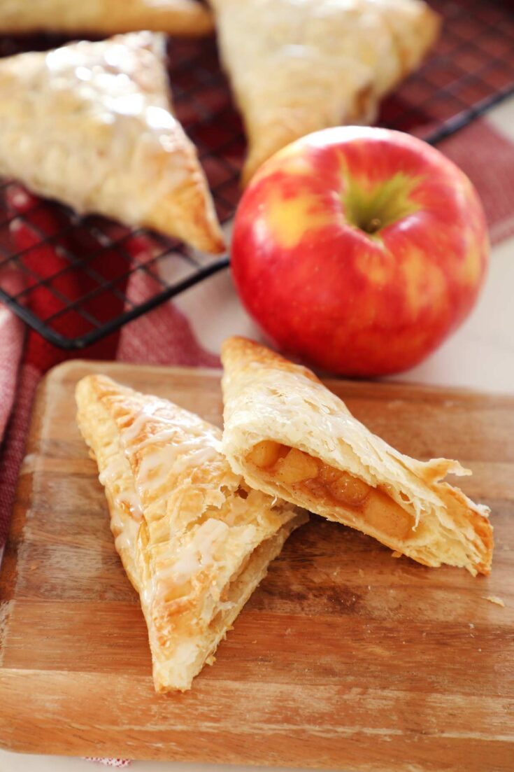 Apple turnovers on a cutting board with an apple and turnovers cooling on a baking rack.