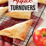 Apple Turnovers resting on cooling rack over red towel and wooden board on kitchen counter, red apple garnish.