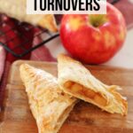 Apple Turnovers with icing resting on cooling rack over red towel and wooden board on kitchen counter, one apple turnover cut in half and red apple garnish.