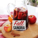 Red Fall Sangria in glass pitcher resting on wooden board and red towel on top of kitchen counter with orange, red apples and cinnamon sticks garnish.