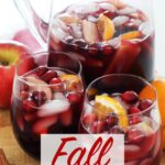 Fall Sangria in glass pitcher and two cups on a wooden board and red towel on top of kitchen counter with orange, red apples, cranberries and cinnamon sticks garnish.
