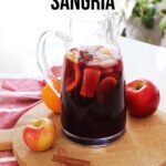 Red Fall Sangria in glass pitcher on a wooden board on top of counter with orange, red apples and cinnamon sticks garnish.