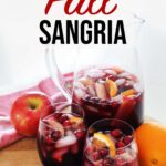 Red Fall Sangria in glass pitcher and cups resting on wooden board and red towel on top of kitchen counter with orange, red apples, cranberries and cinnamon sticks garnish.