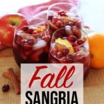 Red Fall Sangria in glass cups resting on wooden board and red towel on top of kitchen counter with orange, red apples, cranberries and cinnamon sticks garnish.