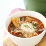 Bowl of lasagna soup on a wooden cutting board with a pot full of soup, green pot in background.