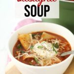 Lasagna Soup in white bowl with spoon resting on wooden board and red towel on top of kitchen counter topped with cheese.