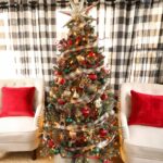 A red and green Christmas tree with plaid ribbon and ornaments with two tan wing backed chair and red pillows.