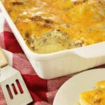 Sausage egg casserole in white casserole dish with one portion plated on red towel on top of kitchen counter.