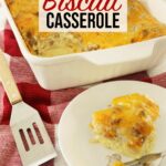 Sausage egg biscuit casserole in white casserole dish with spatula off to the side and one portion on a white plate red towel on top of kitchen counter.