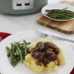 Crock pot beef tips over a bed of mashed potatoes accompanied by some green beans sit on a plate upon the kitchen counter, a full plate of green beans and the crock pot are in the background.